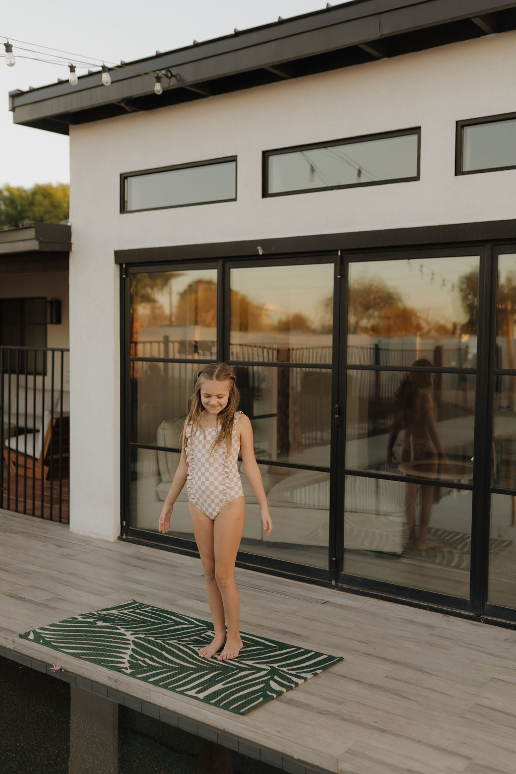 A girl in a forever French baby Girls Sleeveless Swimsuit from the Coastline Collection stands on a deck with a green patterned rug, in front of a modern white house with large glass doors. String lights above her reflect the warm sunset glow.