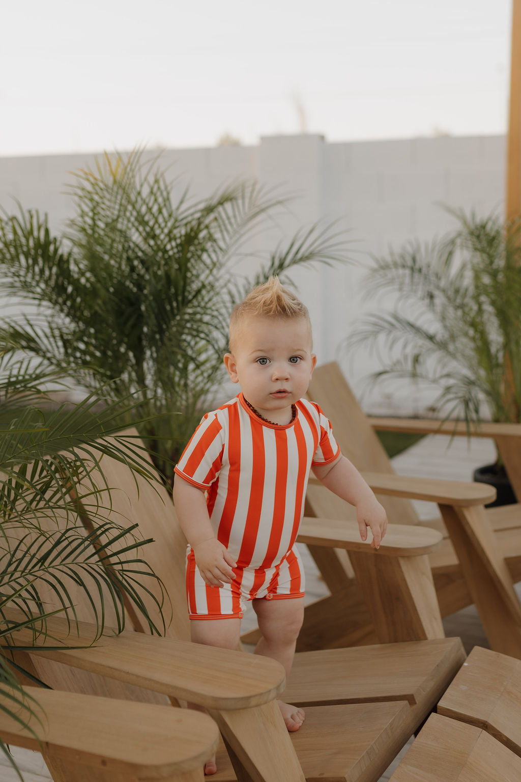 A light-haired toddler in a Forever French Baby orange and white striped Child Shortie Swimsuit, part of the Poolside Collection, stands on a wooden chair amidst furniture and potted plants on a patio. The child looks at the camera, capturing the collection's charm.