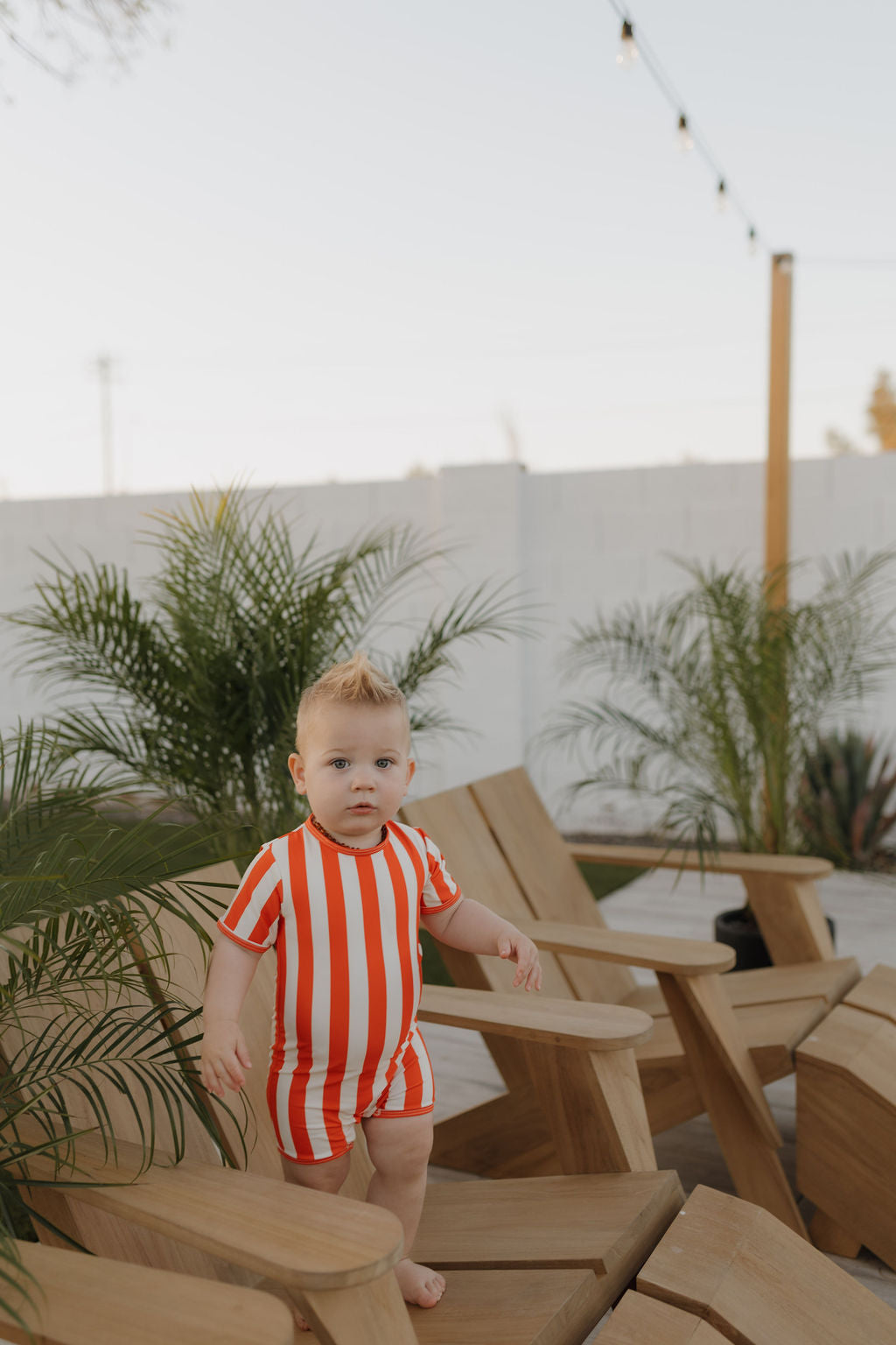 A toddler outdoor on a wooden chair in a red and white Child Shortie Swimsuit from the forever french baby Poolside collection. Potted plants, a white wall, and twinkling string lights create a delightful backdrop under the clear sky.