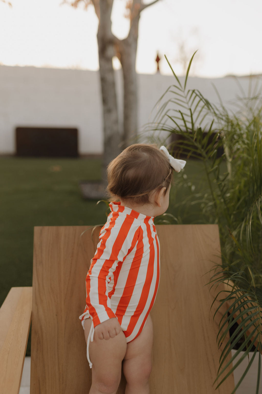 A toddler in a Girls Long Sleeve Swimsuit from "forever french baby" sits on a wooden chair, gazing serenely at the garden's greenery and tree.