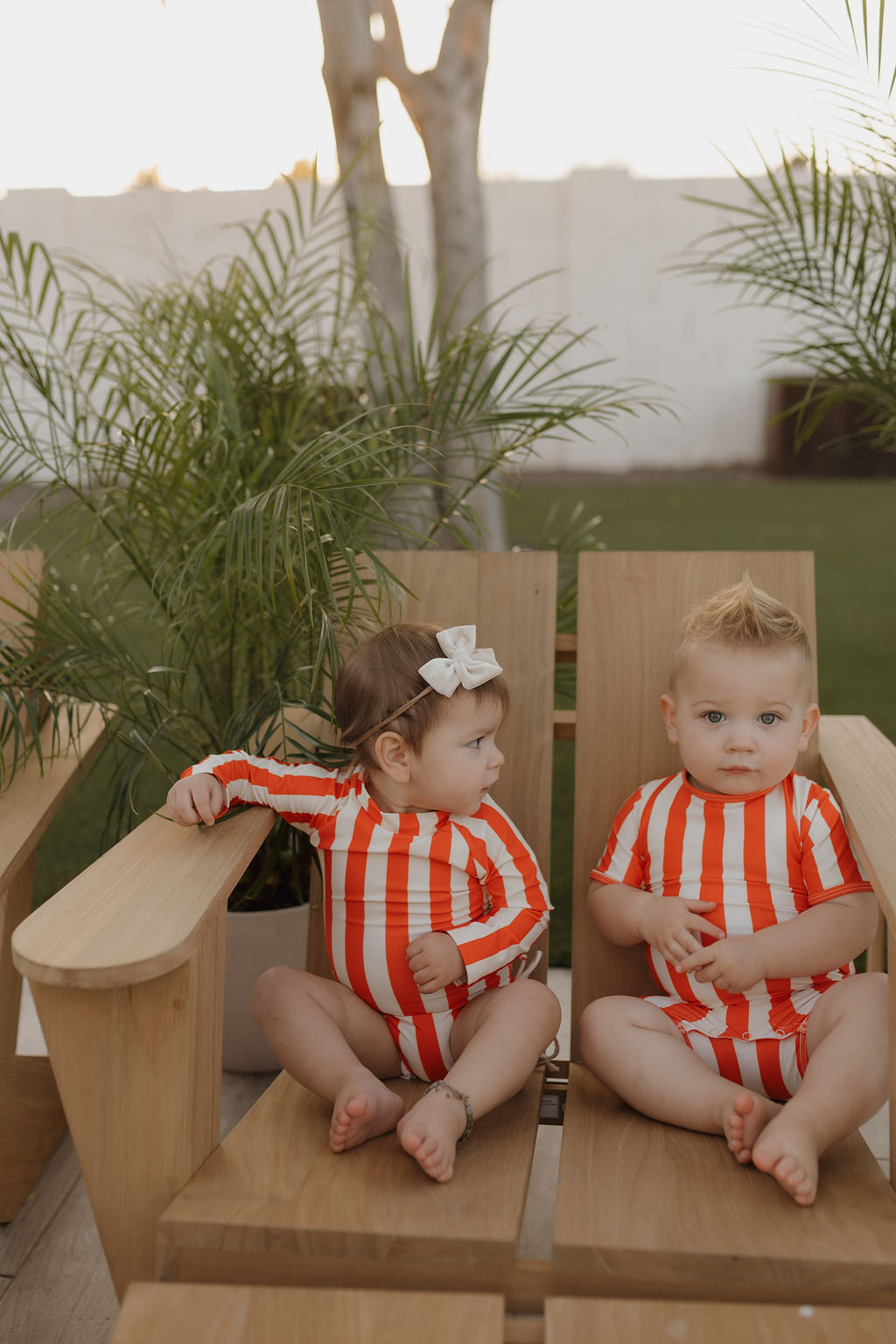 Two toddlers sit outside wearing matching Child Shortie Swimsuits in red and white stripes from the forever french baby brand. One looks to the side while the other gazes forward, framed by lush greenery.