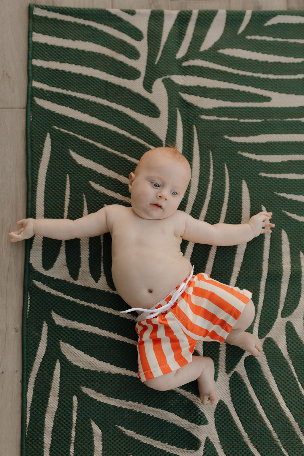 A little one from the forever french baby Poolside collection relaxes on a green and beige leaf-patterned rug wearing orange and white striped Child Boardshorts, adorably spread out on a light wooden floor.