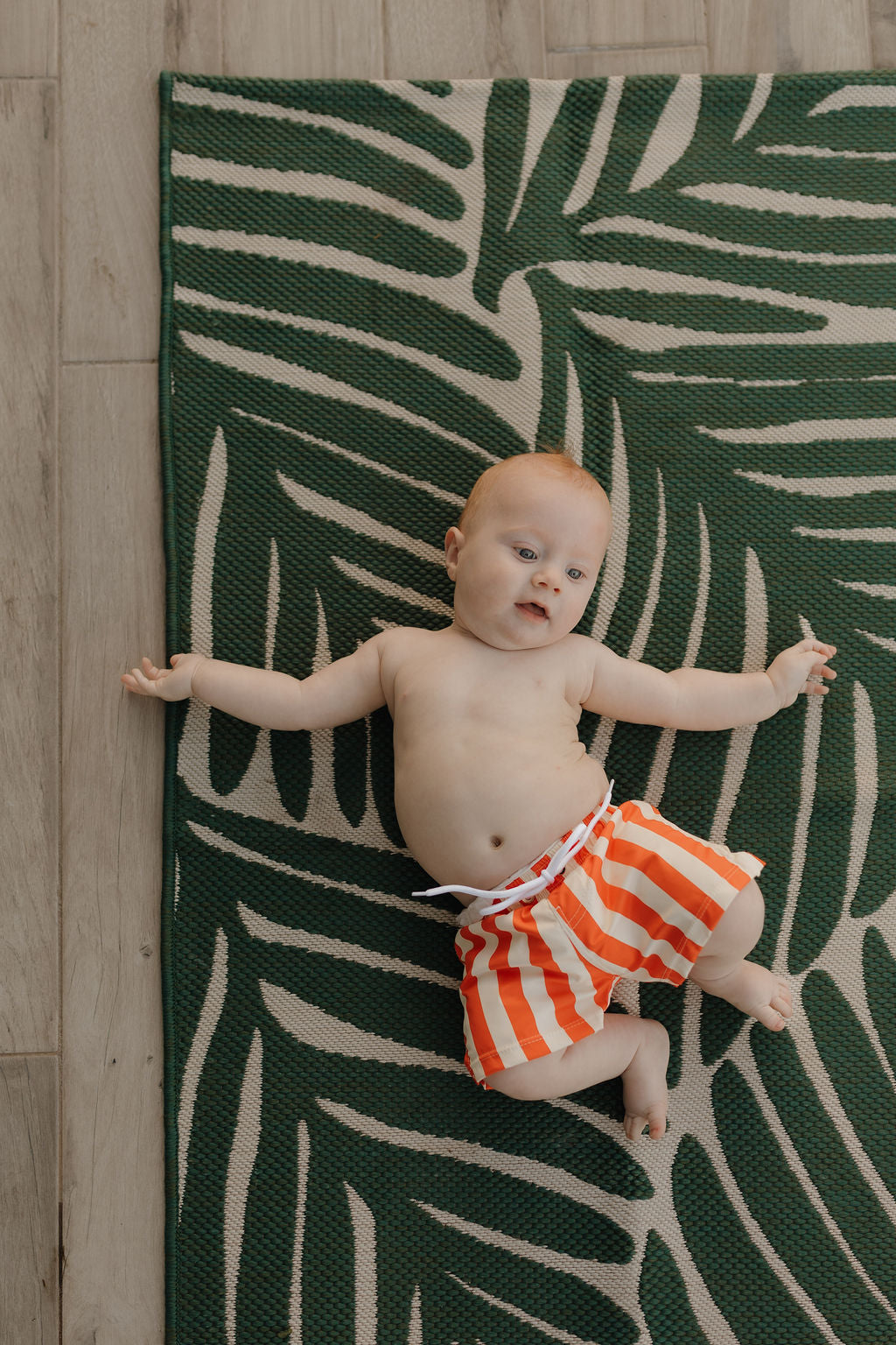 A baby in Forever French Baby's orange/white striped Child Boardshort from the Poolside Collection lies on a leafy green mat atop a wooden floor, joyful with arms and legs spread.