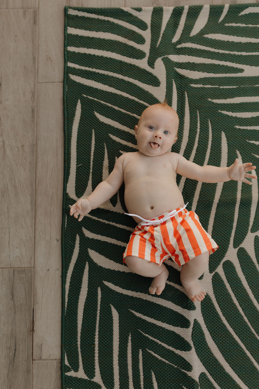 A baby in orange and white Child Boardshorts from Forever French Baby's Poolside collection lies on a leafy-patterned mat, playfully sticking out their tongue. The mat rests on the wooden floor, adding charm to this adorable scene.