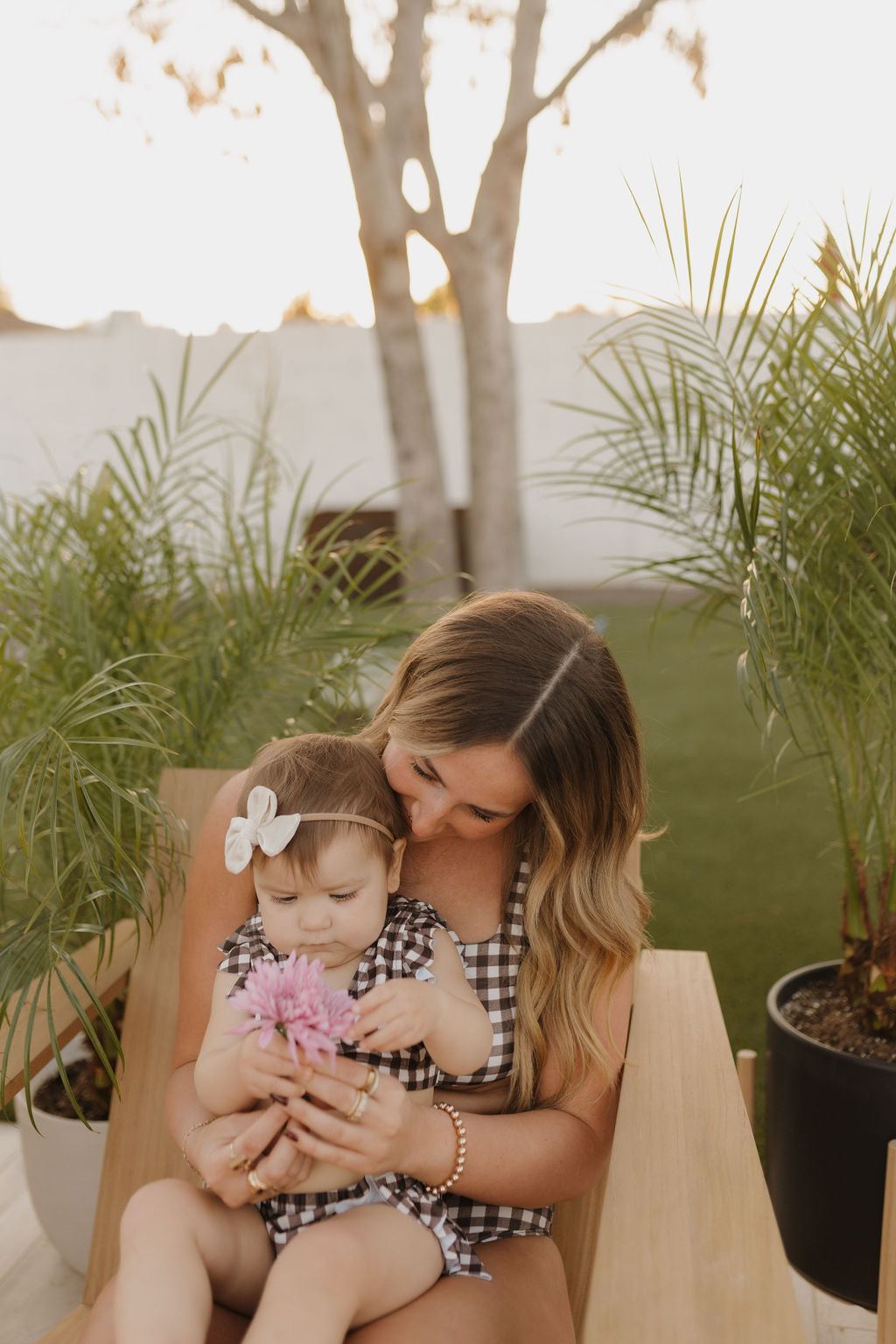 A woman with long hair sits on a wooden chair in the garden holding a toddler dressed in Forever French Baby's Chocolate Gingham swimsuit. Both are in checkered outfits as the toddler gazes at a pink flower, surrounded by palm plants with sunlight filtering through the trees.