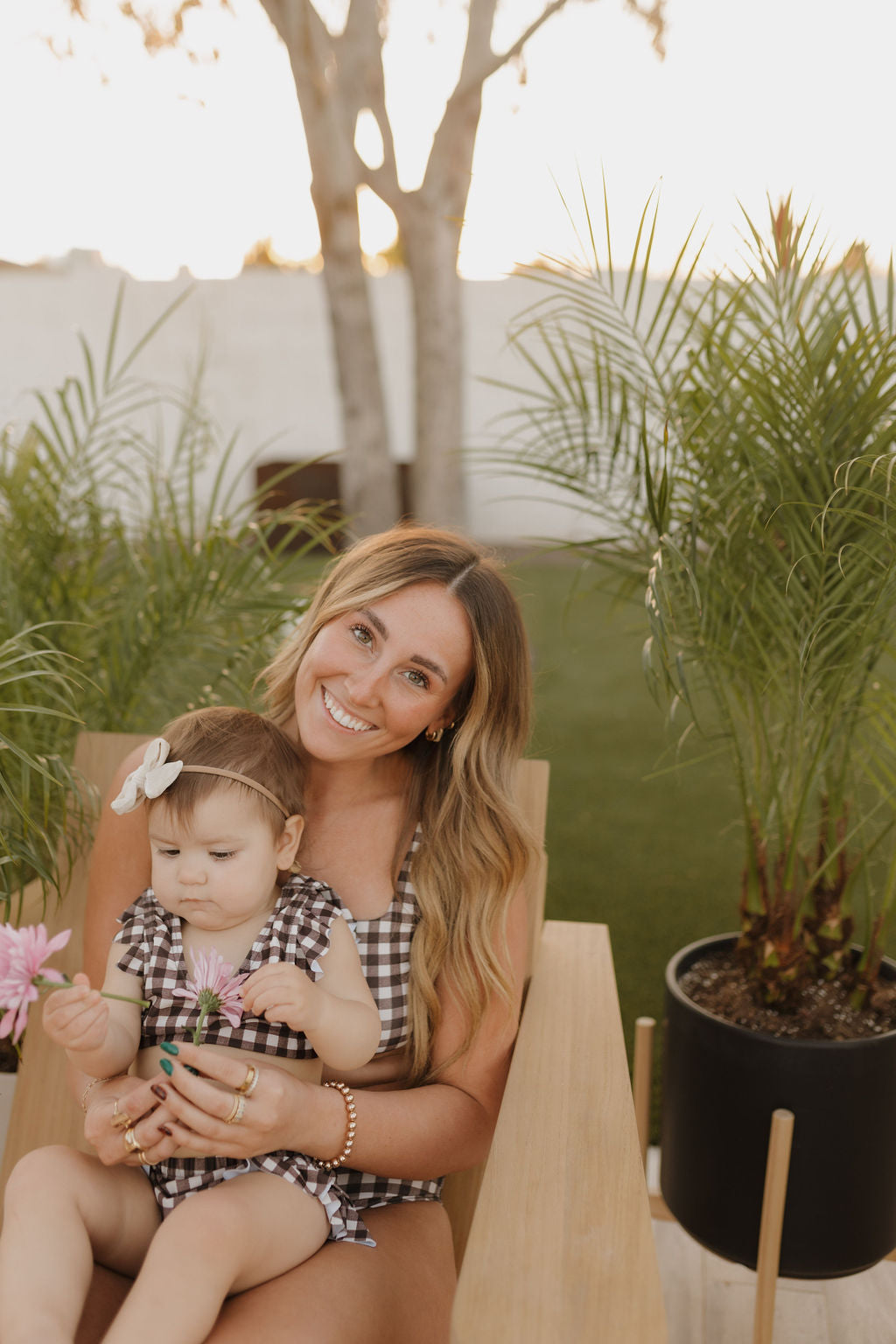 A woman with long hair sits on a wooden chair outdoors, holding a baby from the forever french baby collection wearing the Women's Two Piece Swimsuit in Chocolate Gingham. The baby touches flowers among potted plants, with a tree and sunset behind them. Both are smiling.