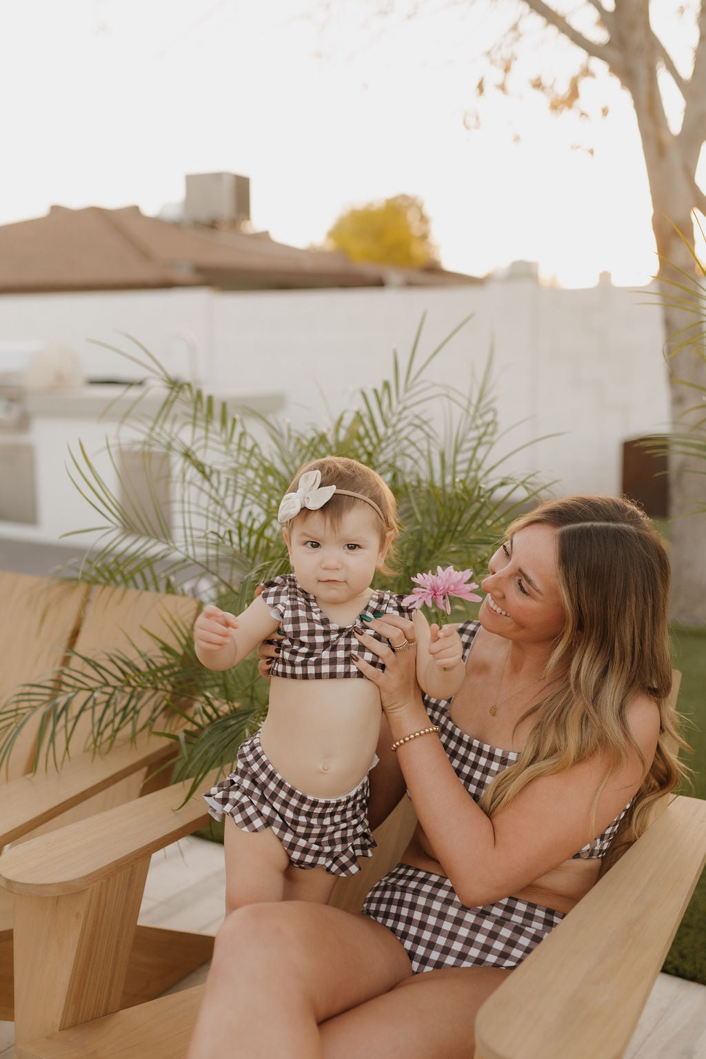 A woman and child, both in forever french baby's Chocolate Gingham swimsuits, sit outdoors on wooden chairs. The child holds a pink flower, and the woman smiles warmly as lush green plants sway gently against a clear sky backdrop.