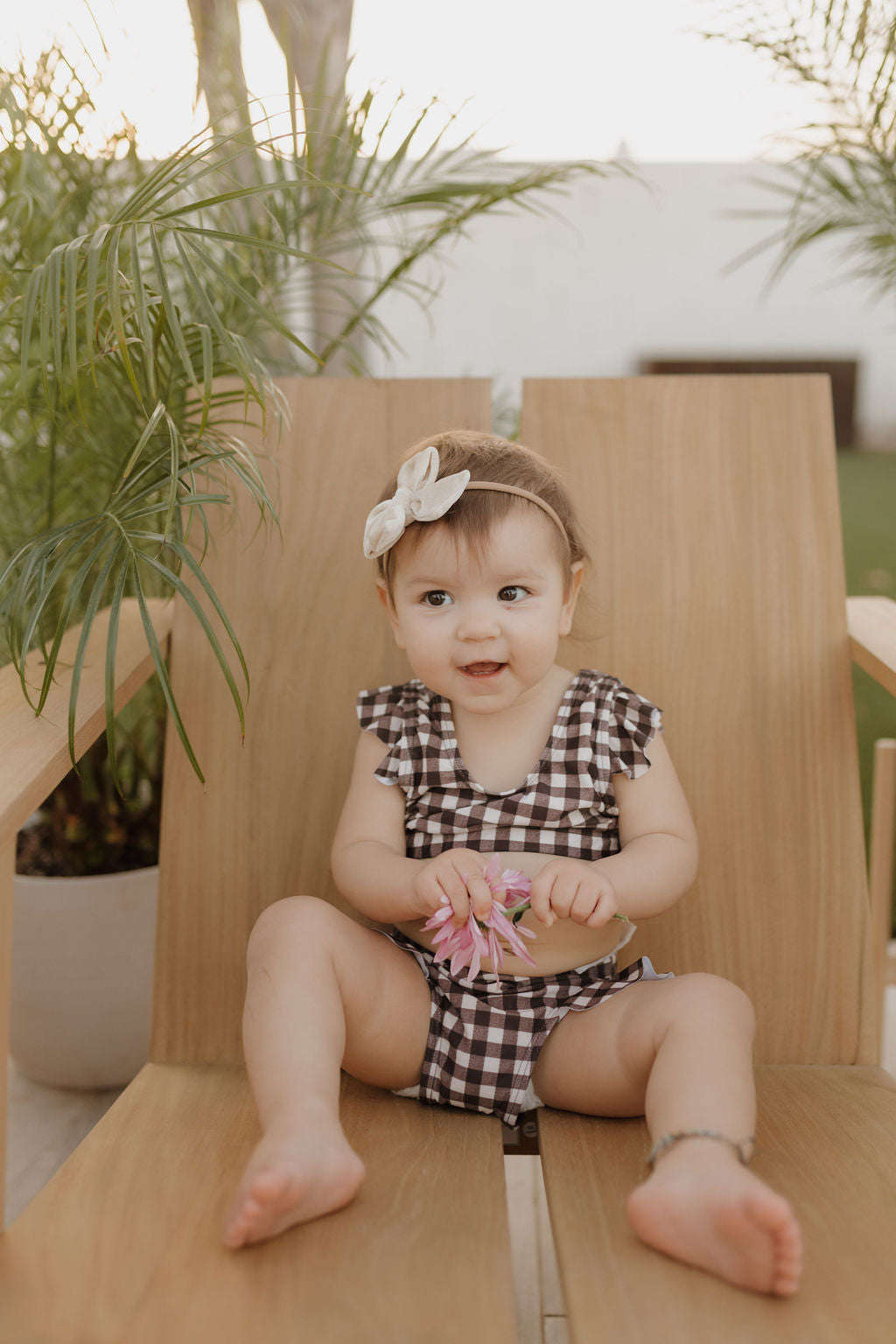 A baby from Forever French Baby sports a checkered outfit from the Chocolate Gingham Collection while seated on a wooden chair outdoors. With ruffled sleeves, she holds a pink flower, wears a beige bow headband, and is surrounded by lush green plants.