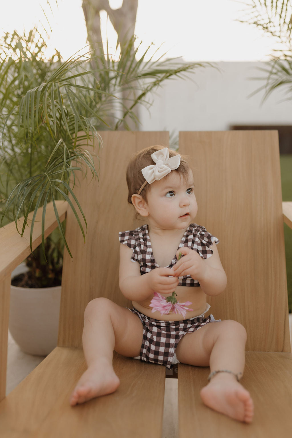 A child from the forever french baby collection sits on a wooden chair, wearing a Girls Two Piece Swimsuit in Chocolate Gingham and a bow headband, holding a pink flower amidst lush greenery that evokes an enchanting outdoor scene.