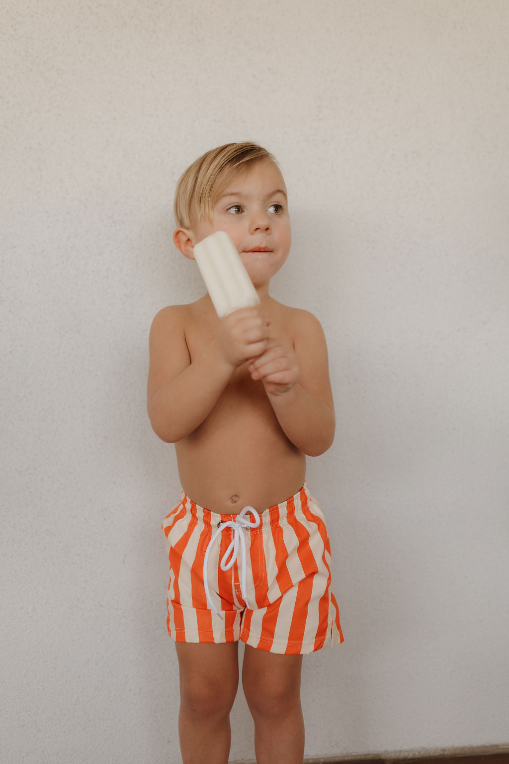 A young child with short, light-colored hair stands indoors against a plain white wall, holding a white popsicle. They're wearing orange and white striped Child Boardshorts from Forever French Baby's Poolside collection.