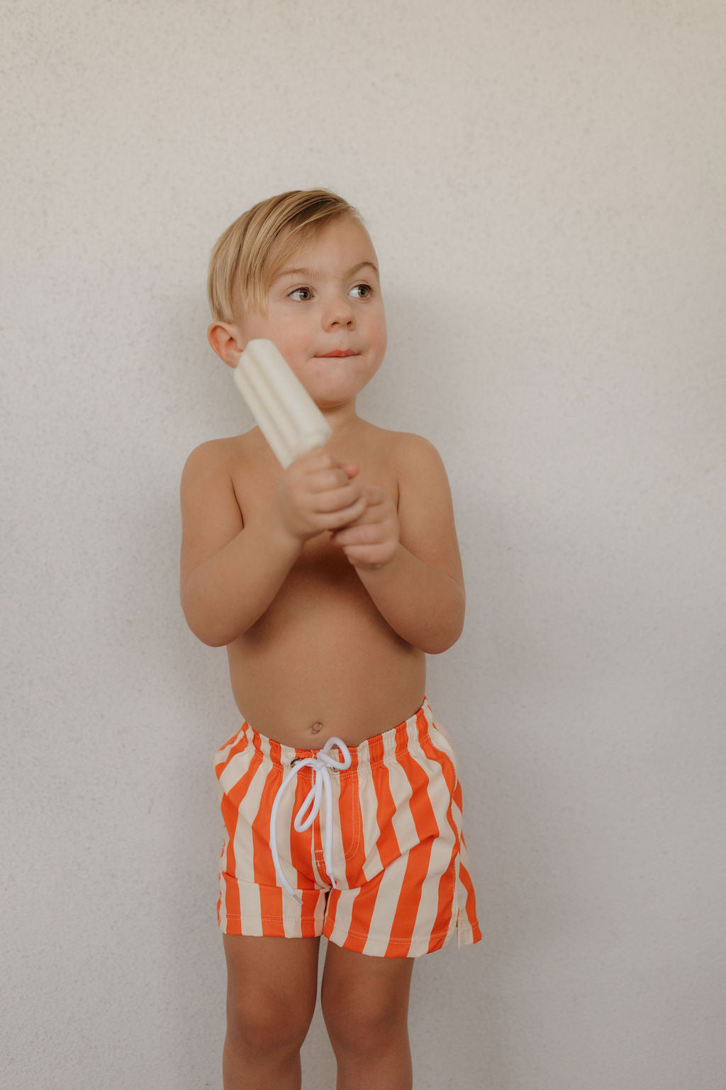 A young child with short blonde hair stands against a plain background, holding a white popsicle. Clad in orange and white striped Child Boardshort | Poolside by forever french baby, the child gazes slightly to the side with a thoughtful expression.