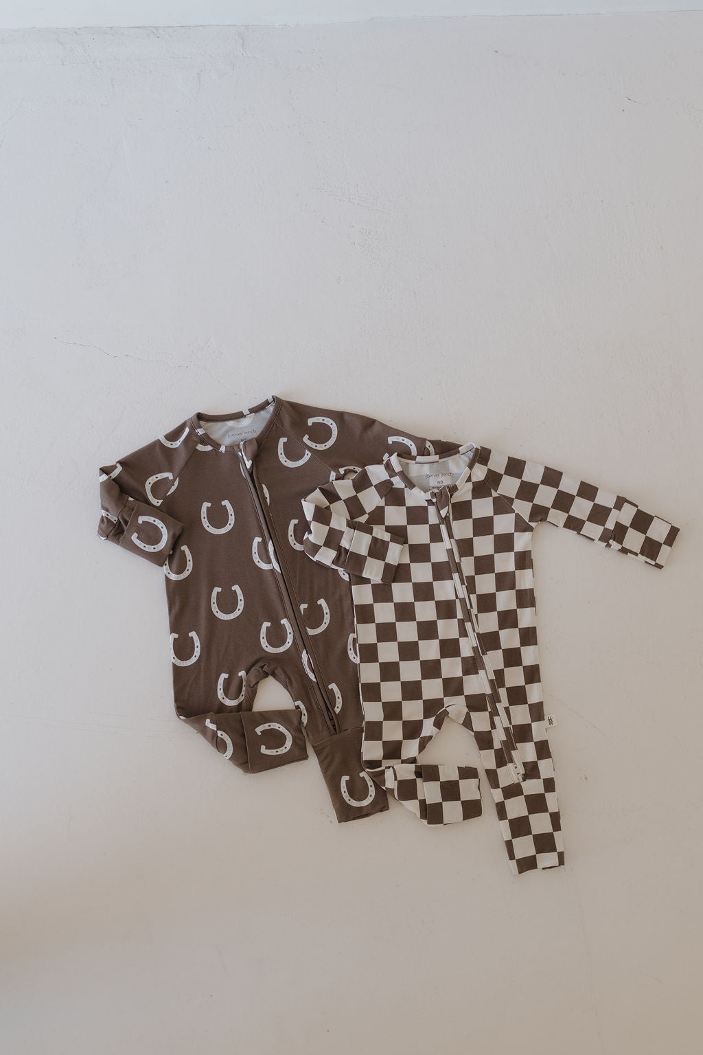 Two Bamboo Zip Pajamas by forever french baby are displayed on a light surface. One features brown with white horseshoe patterns, and the other displays a brown and white checkered design. Both pajamas have front zippers, long sleeves, and are crafted from breathable bamboo fabric.