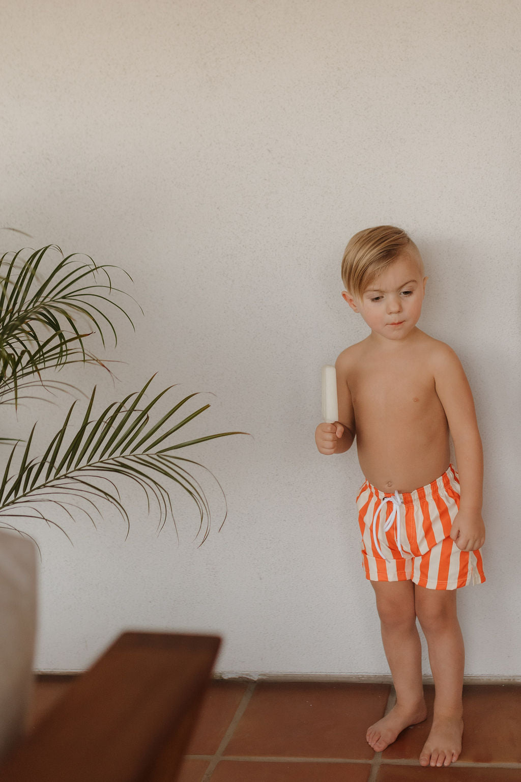 A young child with light hair stands indoors against a white wall, holding a popsicle and wearing Forever French Baby's Child Boardshort from the Poolside collection in orange and white stripes. A potted plant with slender leaves complements the reddish-brown tiled floor on the left.