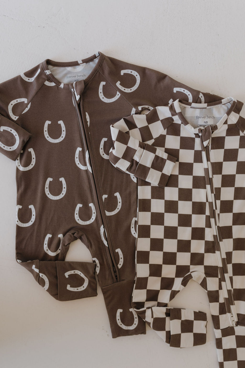 Two Bamboo Zip Pajamas by forever french baby are displayed. One onesie, crafted with hypoallergenic fabric, is brown with white horseshoe patterns. The other features a breathable brown and white checkerboard design. Both have front zippers for easy dressing.