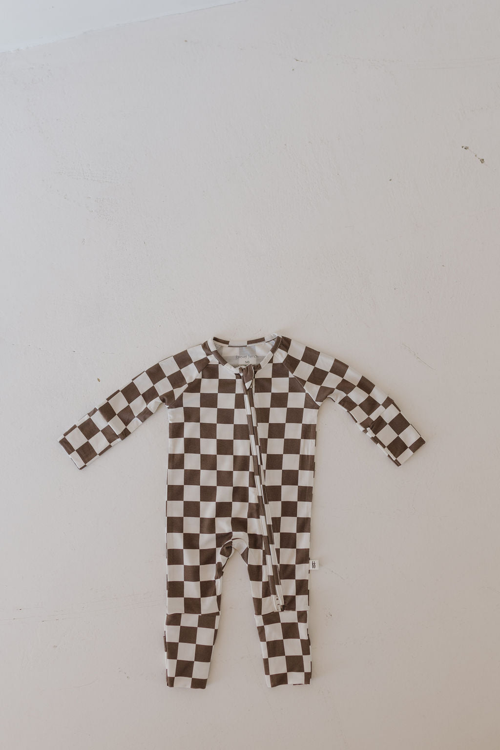 The Bamboo Zip Pajamas | Dutton by forever french baby is a checkered brown and white long-sleeve onesie made from breathable fabric, featuring a front zipper closure, displayed laying flat on a neutral background.