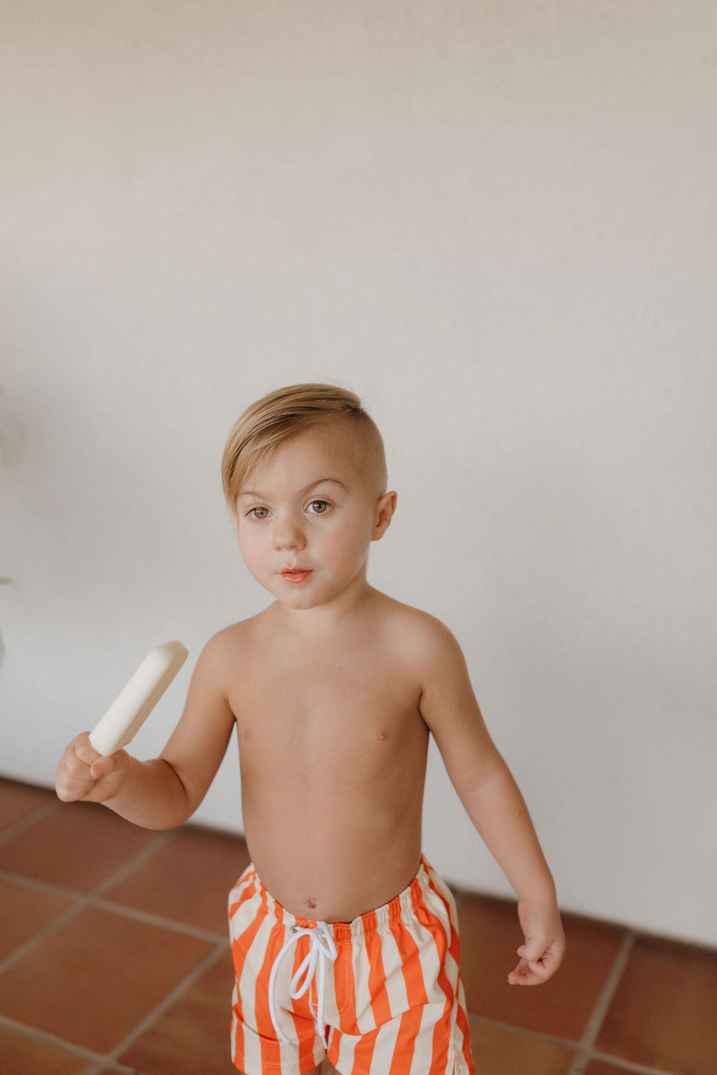 A young child with short hair holds a white popsicle while standing on a brown tiled floor against a neutral backdrop, wearing orange and white striped "Child Boardshort" from the Forever French Baby Poolside collection.