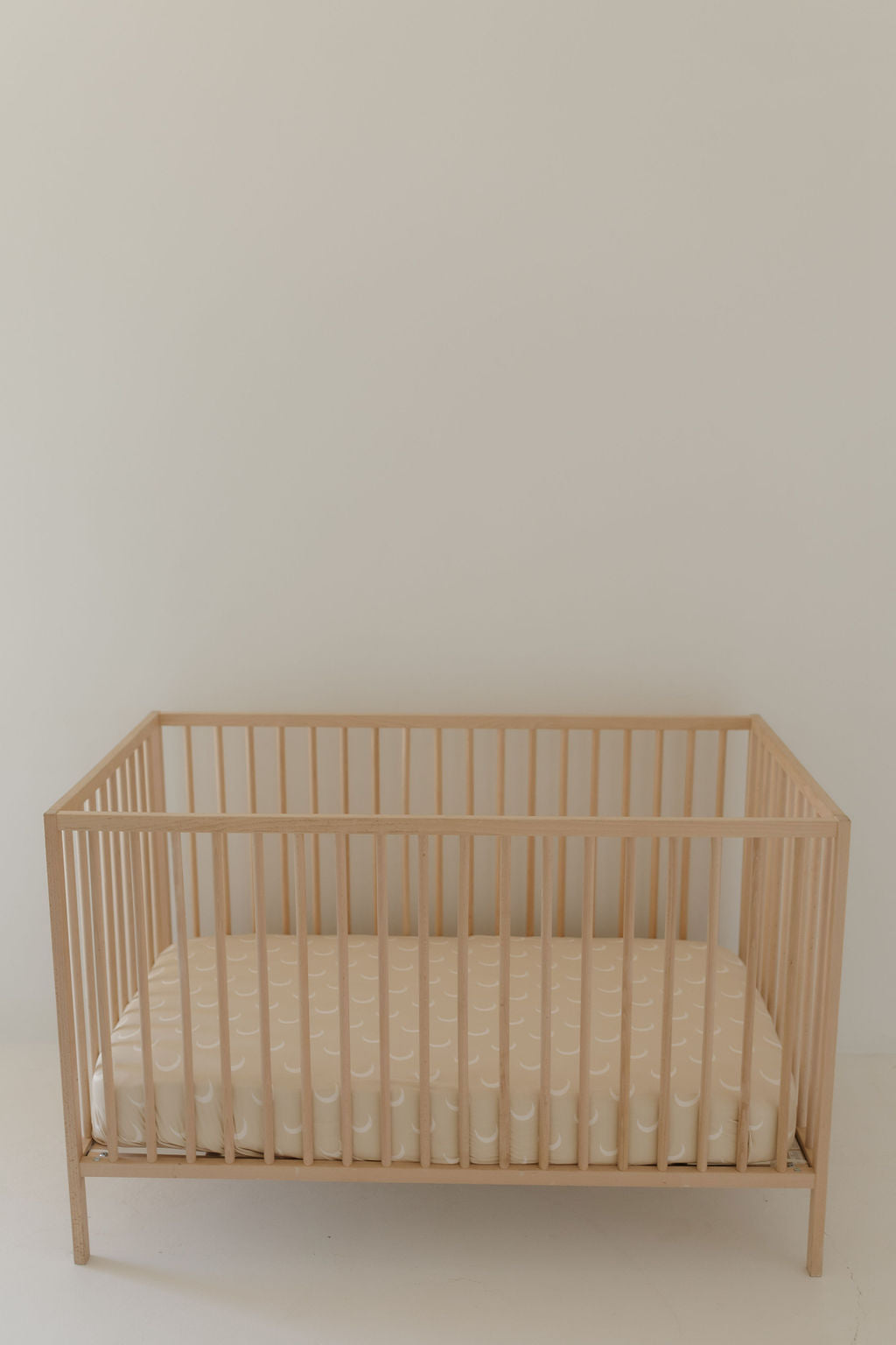 A minimalist wooden baby crib with a Bamboo Crib Sheet | Luna by Forever French Baby and a light-colored mattress featuring a subtle wavy pattern stands against a plain, light-colored wall, creating a simple and serene atmosphere.