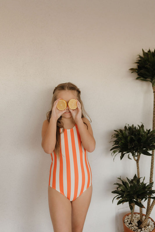 A young girl from the forever French baby Poolside Collection playfully holds lemon halves over her eyes while wearing a Girls Sleeveless Swimsuit. She stands indoors with green potted plants nearby, against a plain wall.