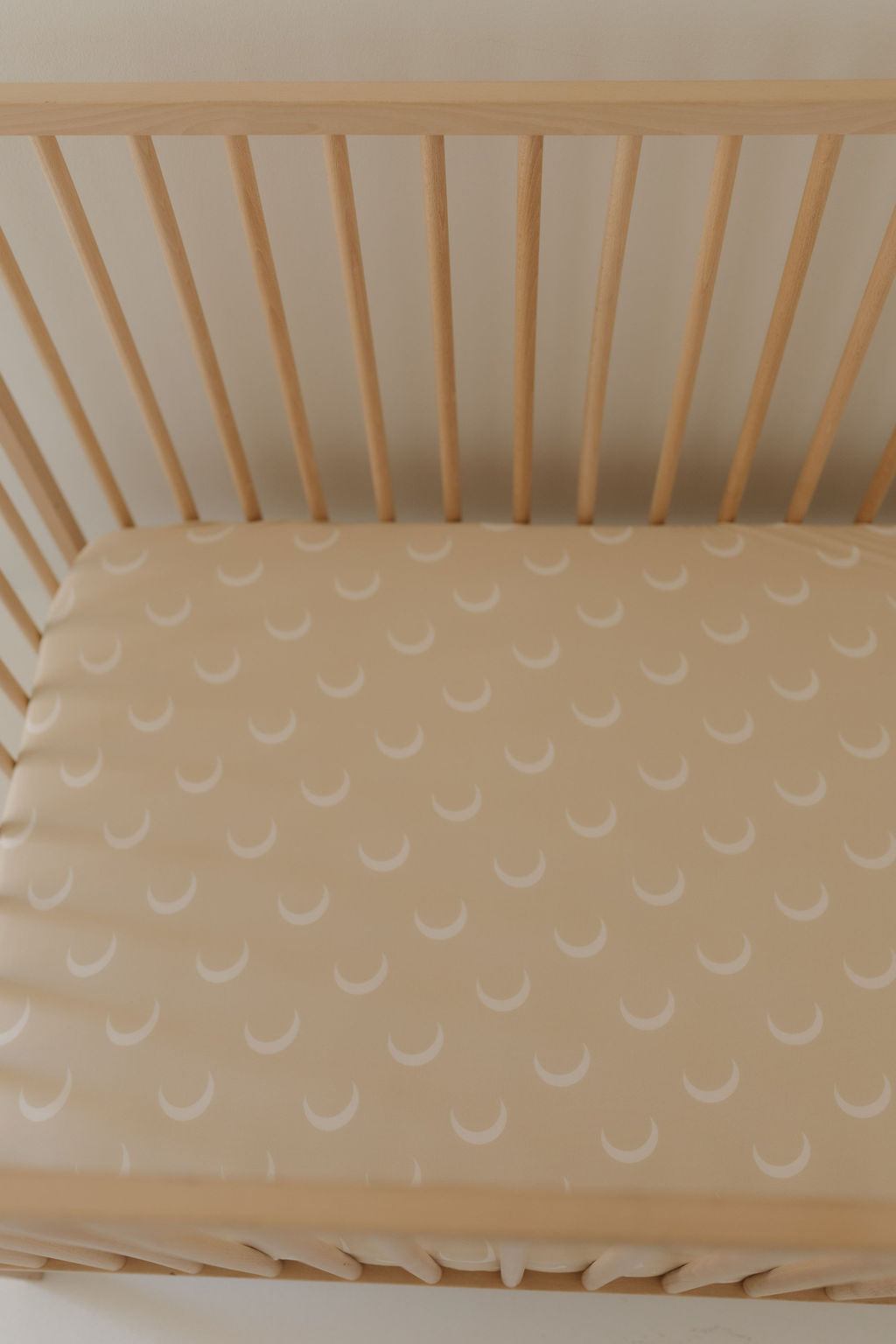 A Forever French Baby bamboo crib sheet, the Luna, features a beige base with white crescent moons and complements the wooden crib's visible slats, creating a serene and inviting nursery setting.