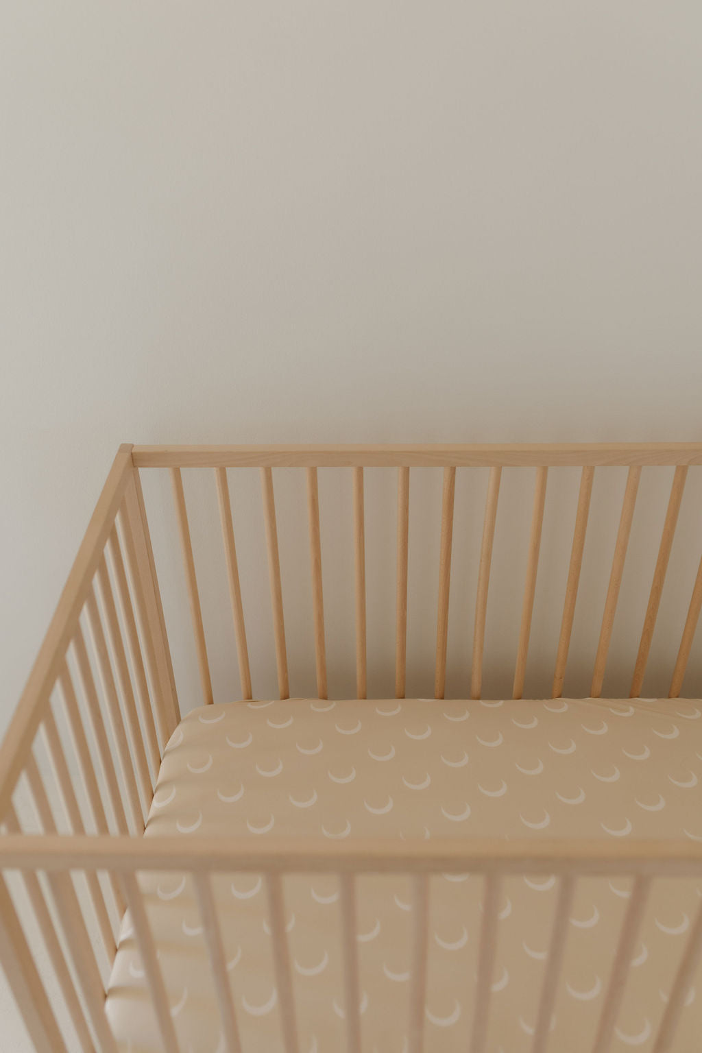 A wooden crib with vertical slats stands against a light-colored wall, its mattress adorned with the Bamboo Crib Sheet | Luna from forever french baby, featuring a pattern of white crescent moons.