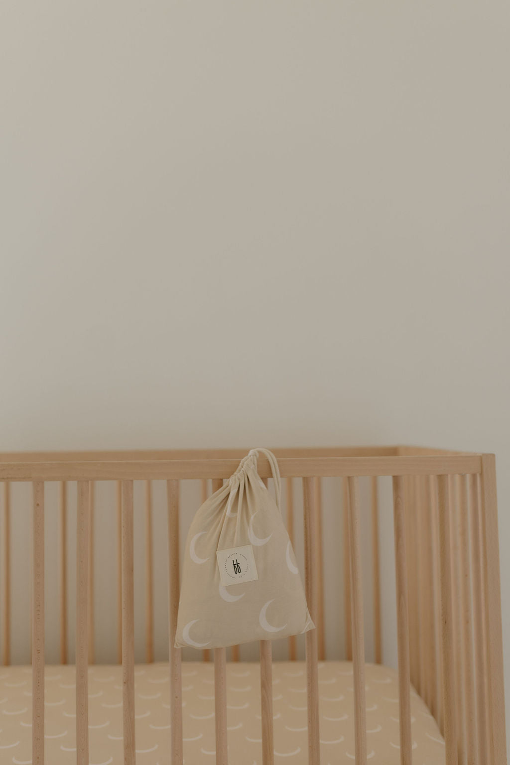 A wooden baby crib with vertical slats showcases a cozy fabric mattress adorned with Forever French Baby's Bamboo Crib Sheet | Luna, featuring a moon pattern on beige. A matching drawstring bag with a white tag hangs from the rail against a plain light-colored wall, enhancing the collection's charm.