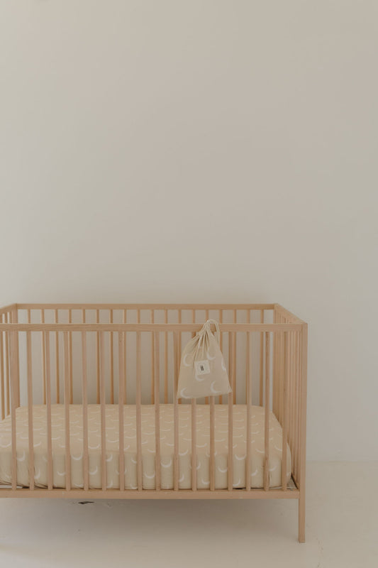 A minimalist nursery highlights a light wooden crib against a plain white wall, featuring the Bamboo Crib Sheet | Luna by forever french baby. A beige-patterned mattress and a small side-hanging bag complete the simple, serene aesthetic, perfect for your Forever French Baby.