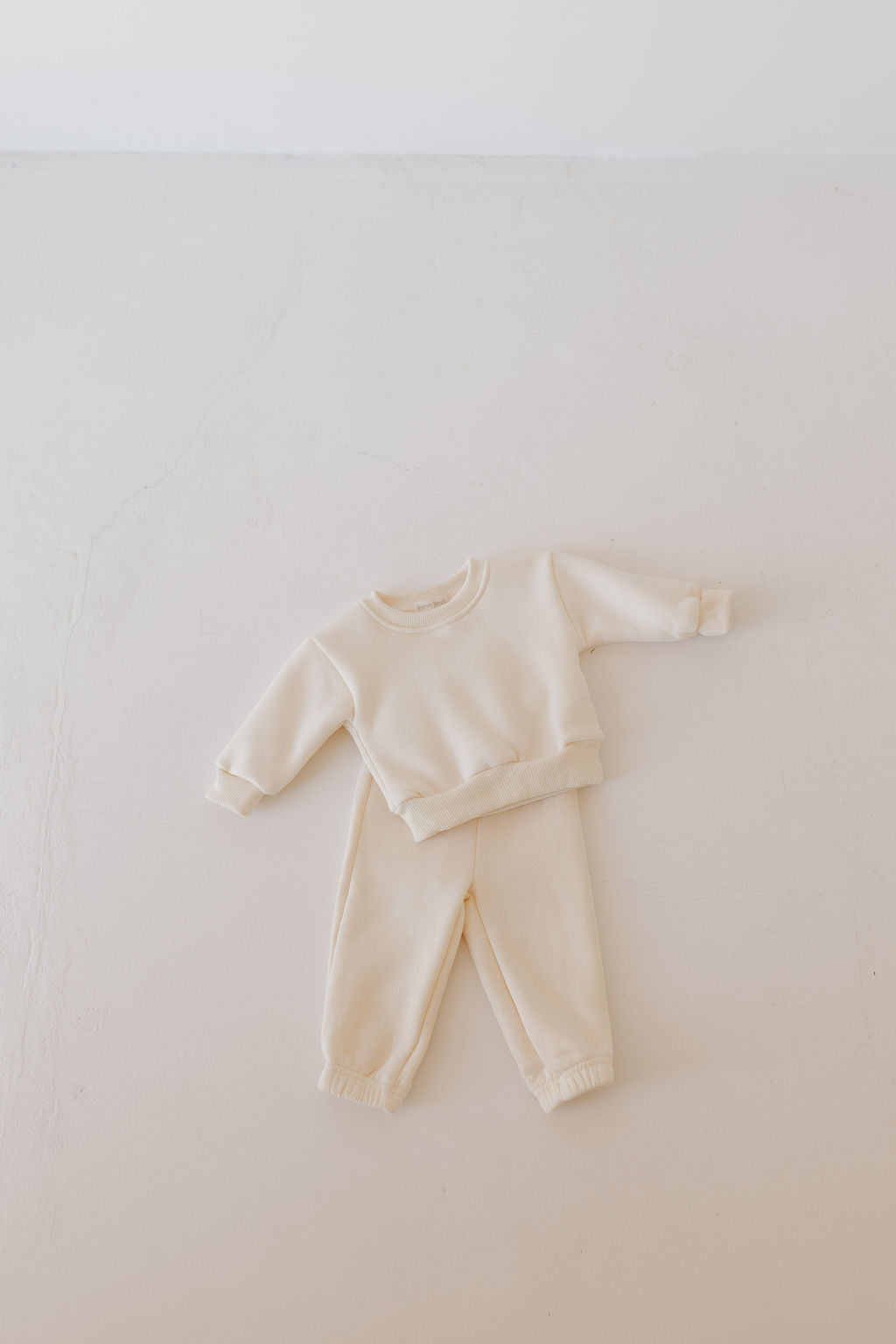 The Sweat Set | Oat from forever french baby is a toddler's outfit, displayed on a smooth, off-white surface. This cream-colored set features an elastic waistband and rolled cuffs, offering a cozy, casual look made from super soft material.