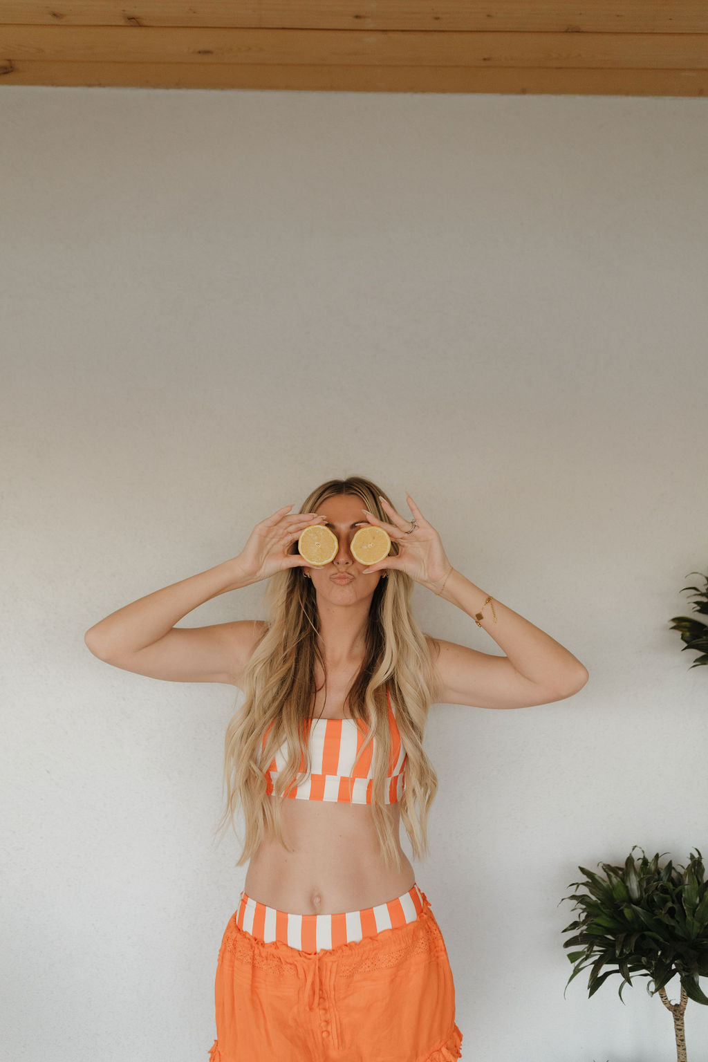 A woman with long blonde hair playfully holds lemon halves over her eyes. Wearing the vibrant orange and white stripes of forever french baby's Women's Two Piece Swimsuit | Poolside, she stands against a plain wall with green plants nearby, exuding summer fun sans her high-waisted bikini bottom.