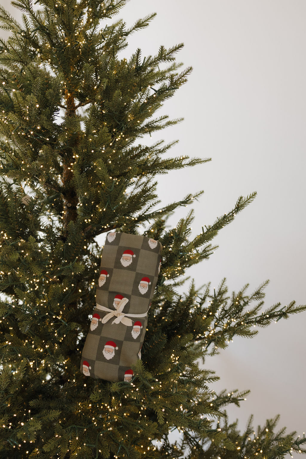 A Muslin Swaddle | Santa Checkerboard (Preview) by forever french baby, wrapped with a white ribbon, sits among Christmas tree branches with string lights, set against a plain white background.