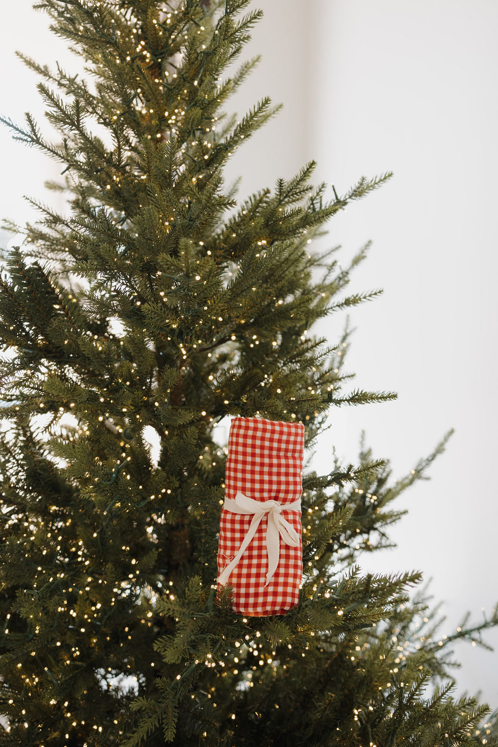 A softly lit Christmas tree glows with white lights, featuring the forever french baby Muslin Swaddle | Red Gingham—wrapped like a gift with a white ribbon—hanging delicately from a branch. The background is uncluttered.
