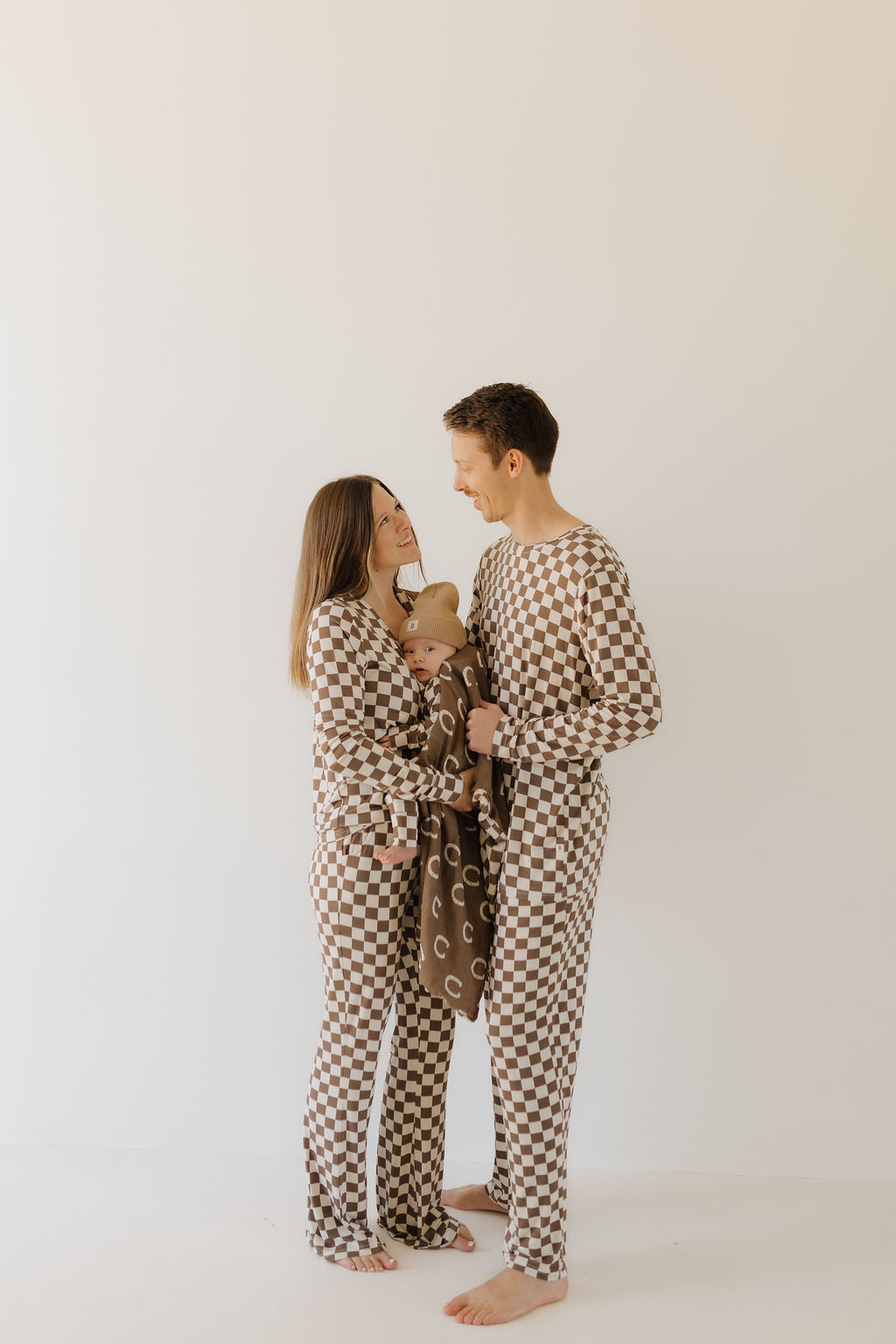 A couple stands barefoot in matching forever french baby Bamboo Zip Pajamas | Dutton, holding an infant wrapped in a patterned blanket. They smile at each other against a plain light background, enjoying the breathable fabric that promises comfort for their little family.