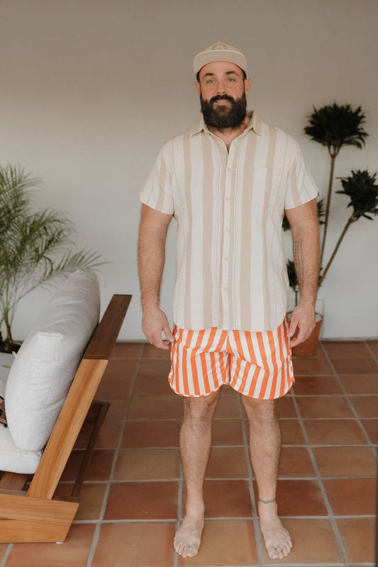 A bearded man from Forever French Baby wears a beige cap and cream-striped shirt with Men's Boardshort in orange stripe from the Poolside collection, standing on a tiled floor. A beige sofa and potted plants are behind him.