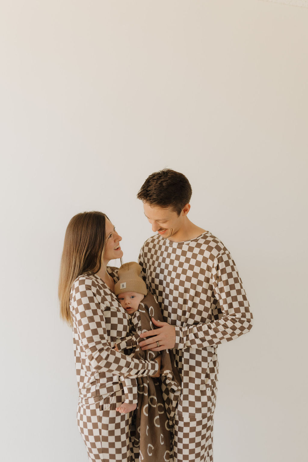 A family of three, dressed in forever french baby's bamboo zip pajamas in a brown and white checkered pattern, stands together. The woman holds a baby wrapped in a designed brown blanket. The smiling man gently touches the baby's head against an off-white background.