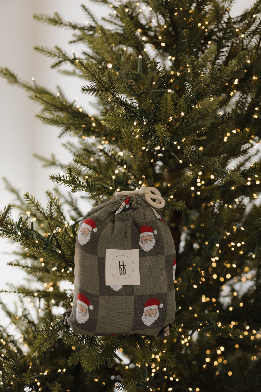 A forever french baby Muslin Crib Sheet in Santa Checkerboard print adds cozy, festive charm to your nursery—just like a Santa-patterned drawstring gift bag on a warmly lit Christmas tree.