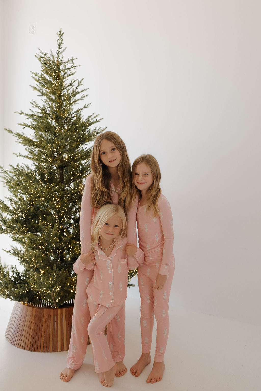Three girls smile and pose barefoot beside a white-lit Christmas tree in a bright, minimal room, wearing matching forever french baby Bamboo Two Piece Pajamas | Nutcracker.