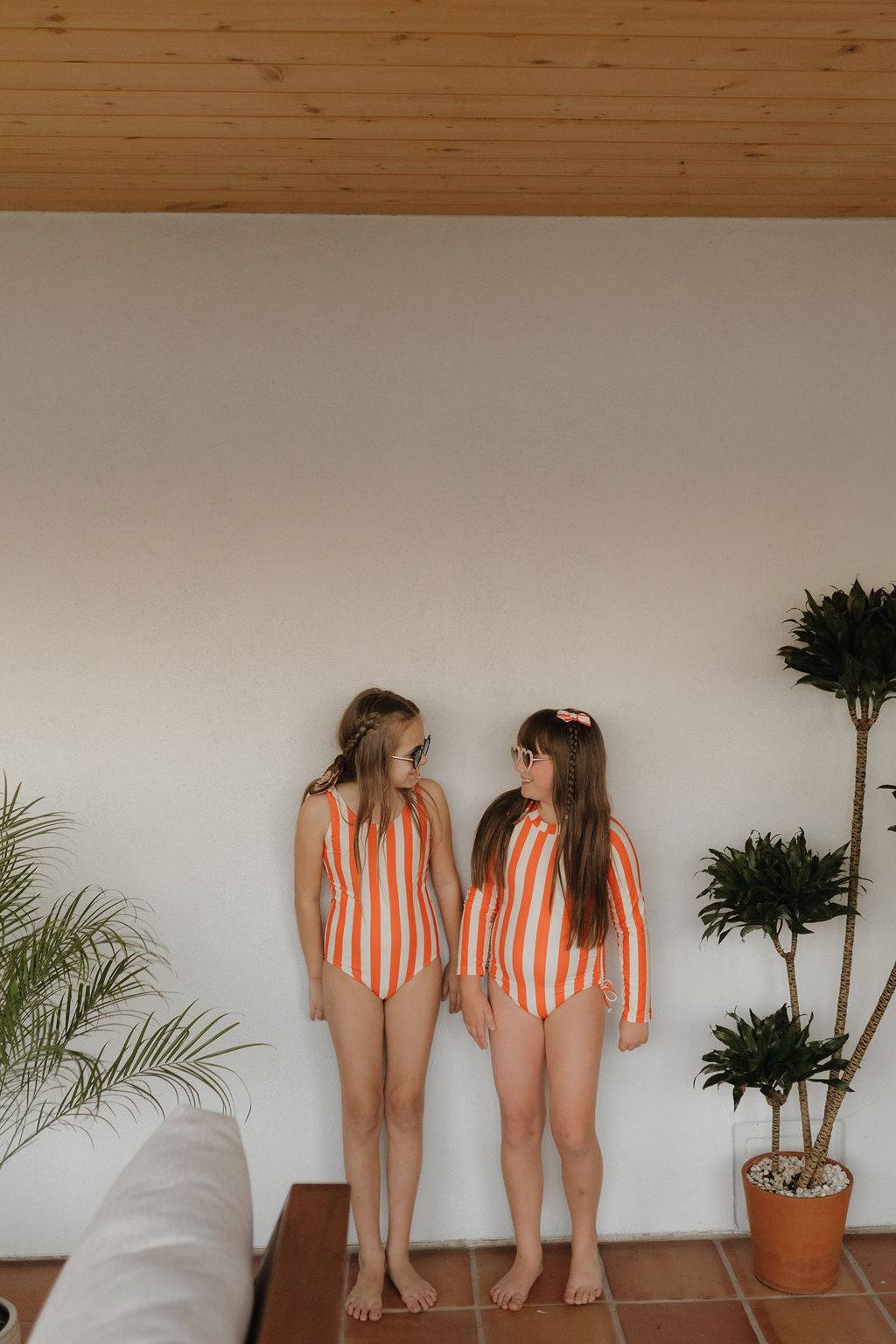 Two kids, wearing matching Girls Sleeveless Swimsuits from the Forever French Baby Poolside Collection, smile at each other indoors. A potted plant is to their right against a white wall, with part of a sofa visible on the left.