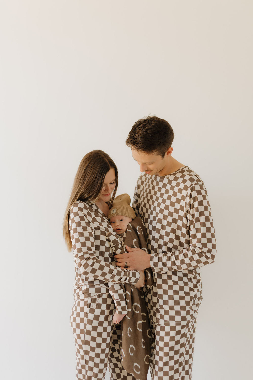 A family of three, dressed in matching breathable fabric outfits from Forever French Baby's Bamboo Zip Pajamas | Dutton, stands against a white background. The parents gently hold their baby, cozy in a brown blanket with a hat.