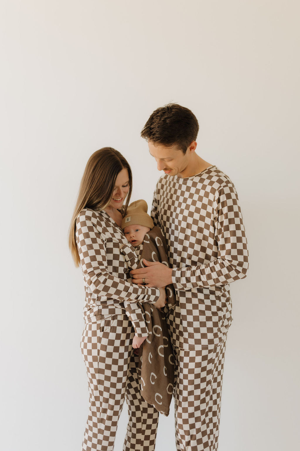 A couple in brown and white checkered outfits lovingly holds a baby wrapped in a brown blanket and tan beanie. The baby is cozy in forever french baby's Bamboo Zip Pajamas | Dutton, crafted from breathable fabric. They stand against a plain white background.