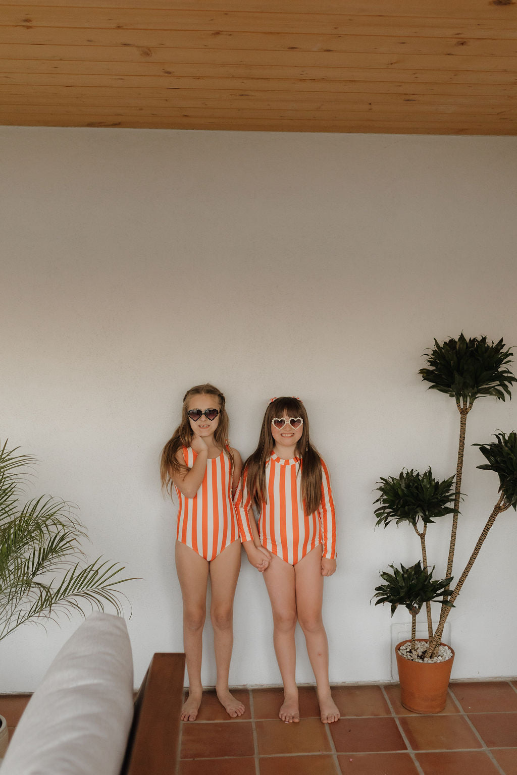 Two kids in Forever French Baby's Poolside Collection striped swimsuits stand hand-in-hand indoors, radiating summer vibes, framed by a white wall, potted plants, and a tiled floor.