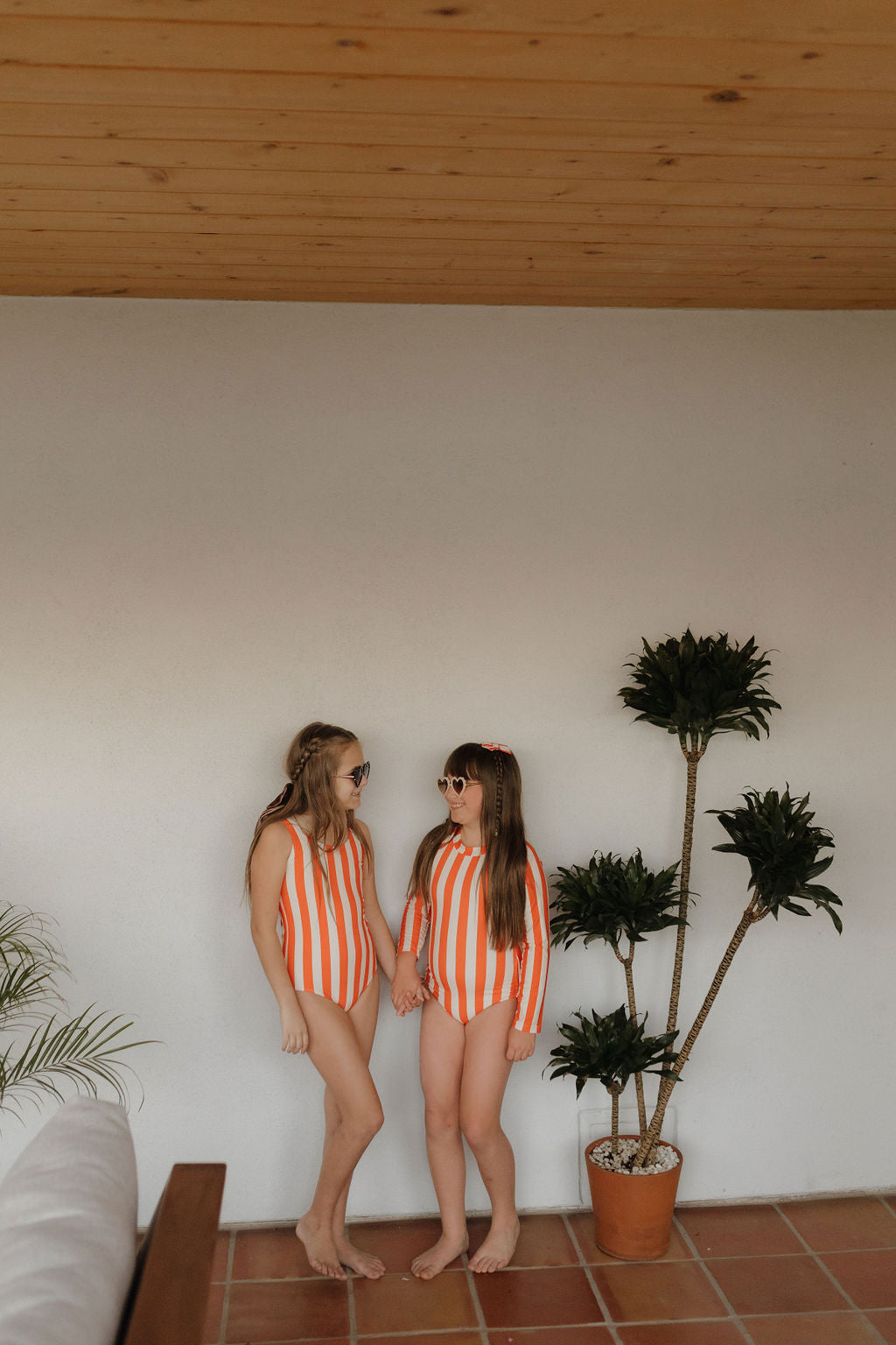 Two children wear matching Girls Sleeveless Swimsuits from the forever French baby Poolside Collection with chic sunglasses. They stand hand in hand on a tiled floor with a potted plant nearby and a wooden ceiling above, capturing the essence of summer fun.