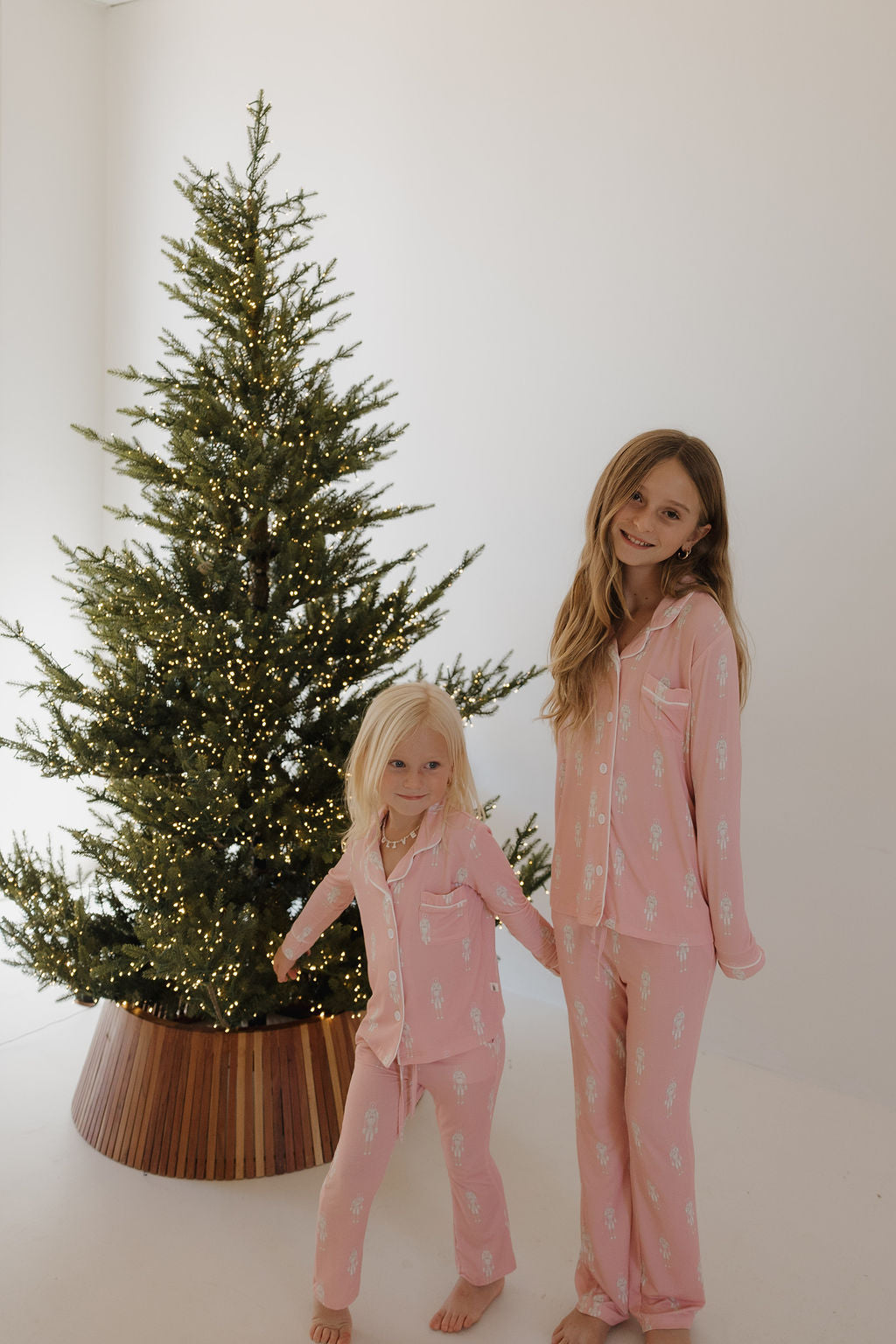 Two young girls in matching forever french baby Pre-Teen Flare Bamboo Pajamas | Nutcracker smile beside a decorated Christmas tree with white lights in a bright, simple room.