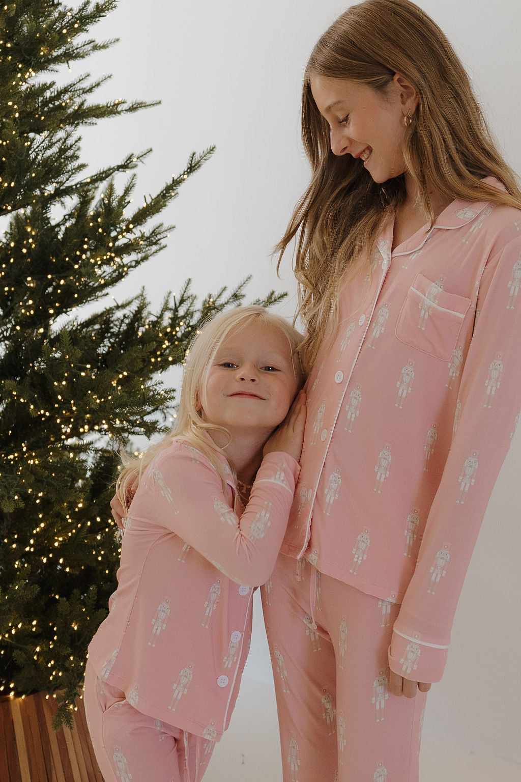 Two smiling children in forever french baby Pre-Teen Flare Bamboo Pajamas | Nutcracker stand by a festive Christmas tree. The younger hugs the older, showing the comfort and hypo-allergenic quality of these holiday pajamas.