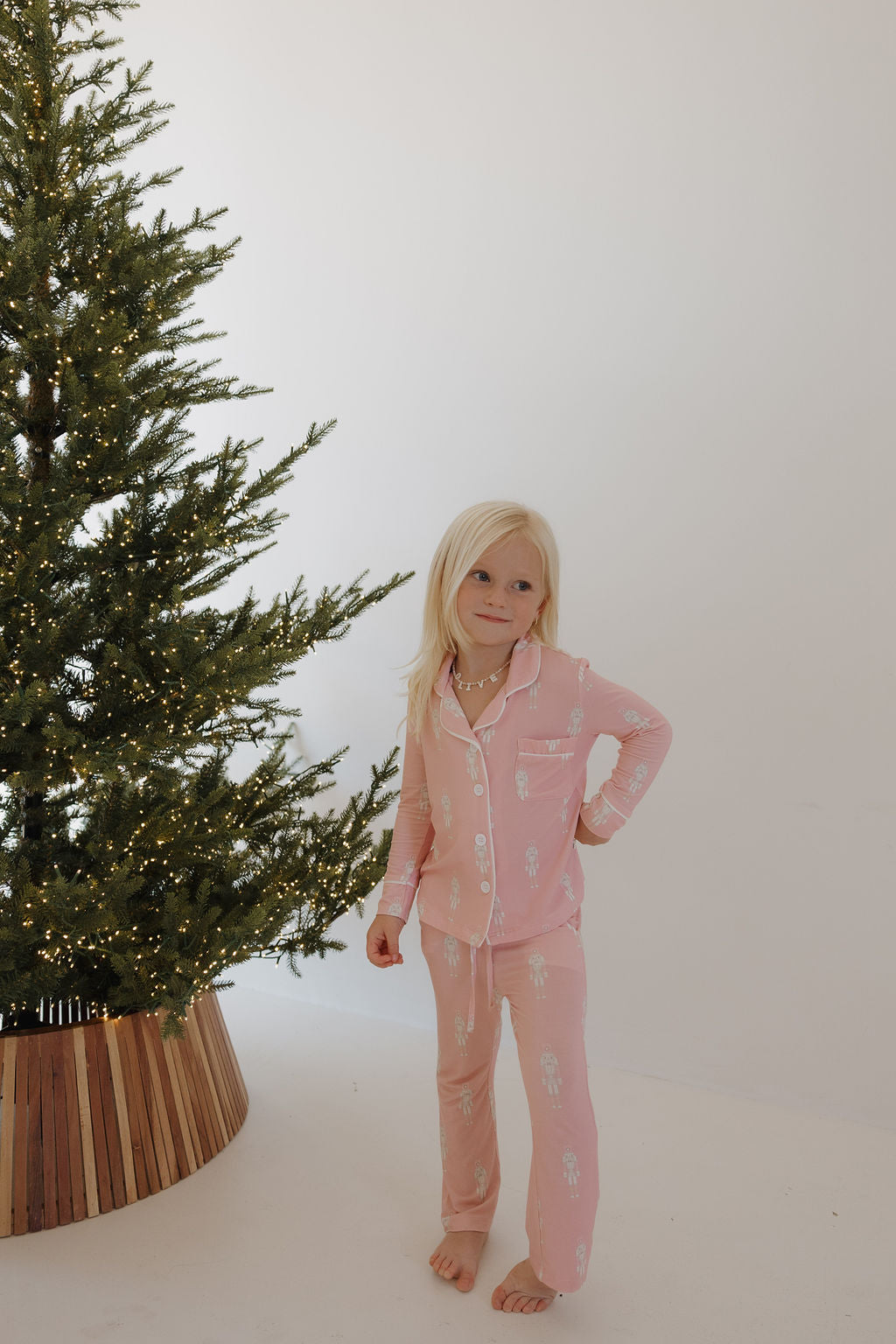 A young girl with long blonde hair stands barefoot in forever french baby’s Long Sleeve Toddler Bamboo Pajamas | Nutcracker beside a decorated Christmas tree with white lights, against a plain white background.