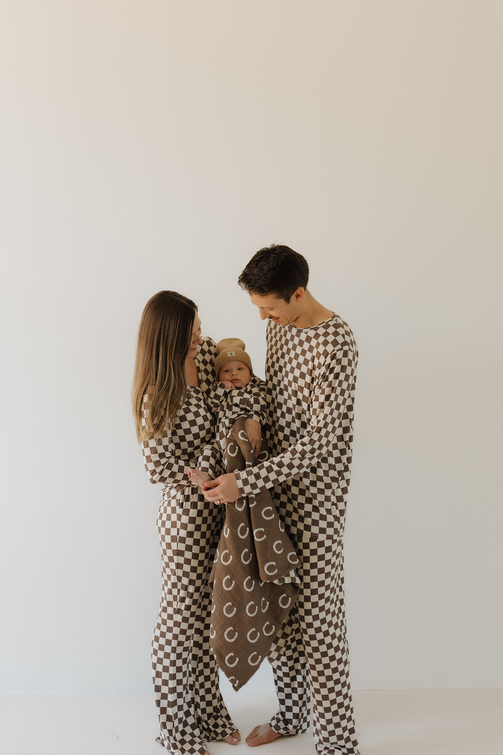 A family of three poses together. The parents wear matching brown and cream checkered pajamas, identified as the Men's Bamboo Pajama from Dutton by forever french baby. Their baby is wrapped in a cozy blanket featuring letter "C" patterns, all against a plain background.