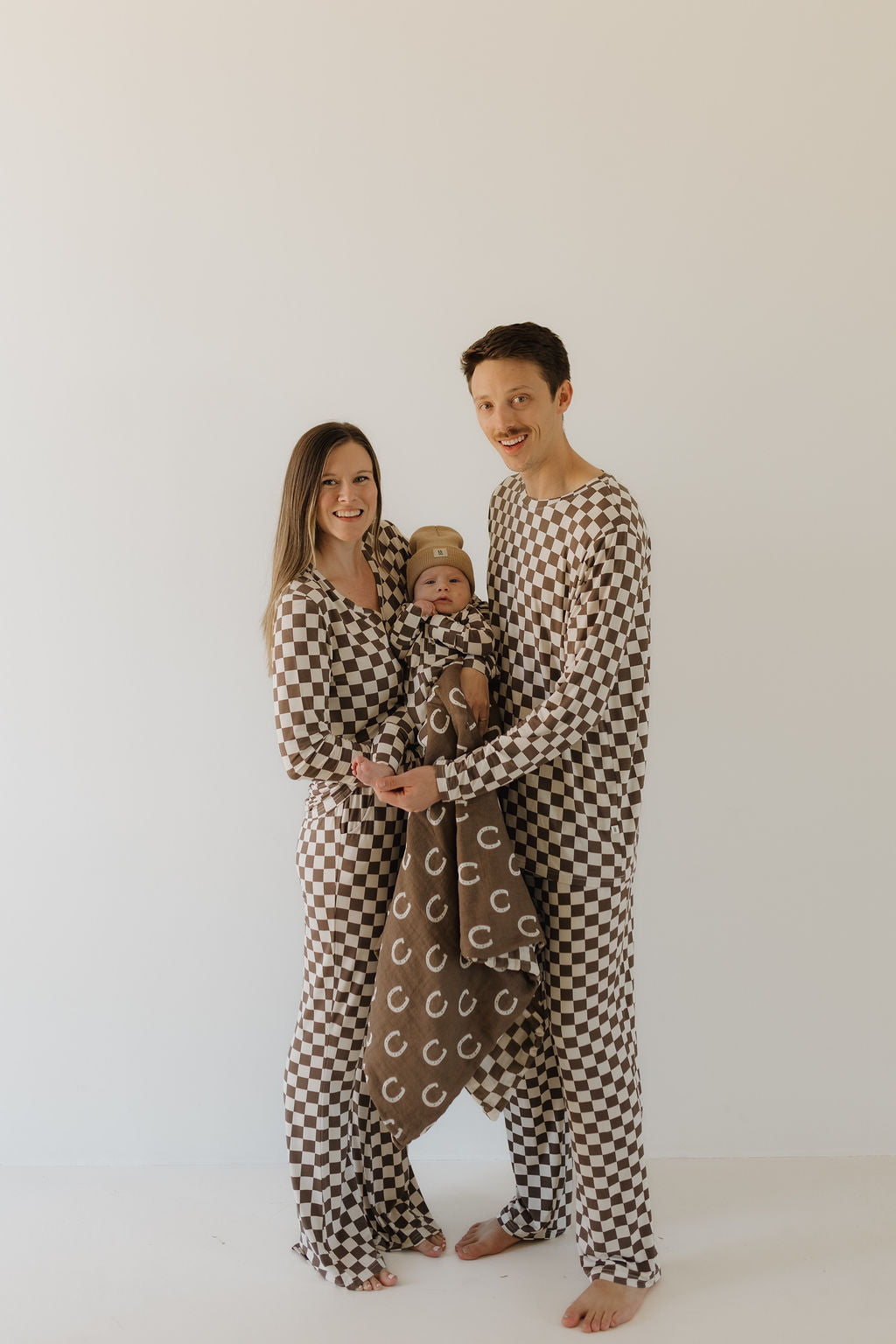 A smiling family of three wears matching Bamboo Zip Pajamas by Forever French Baby against a plain background. The parents hold their baby wrapped in a cozy, matching patterned blanket.