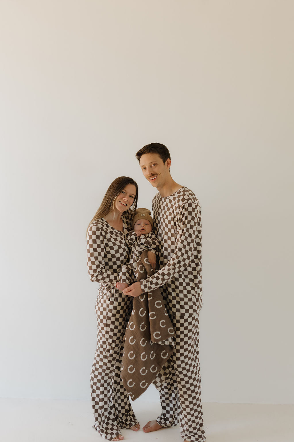 A family of three, dressed in matching forever french baby Bamboo Zip Pajamas | Dutton with a brown and white checkered design, smiles against a white background. The parents hold their baby wrapped in a soft brown blanket featuring a white pattern.