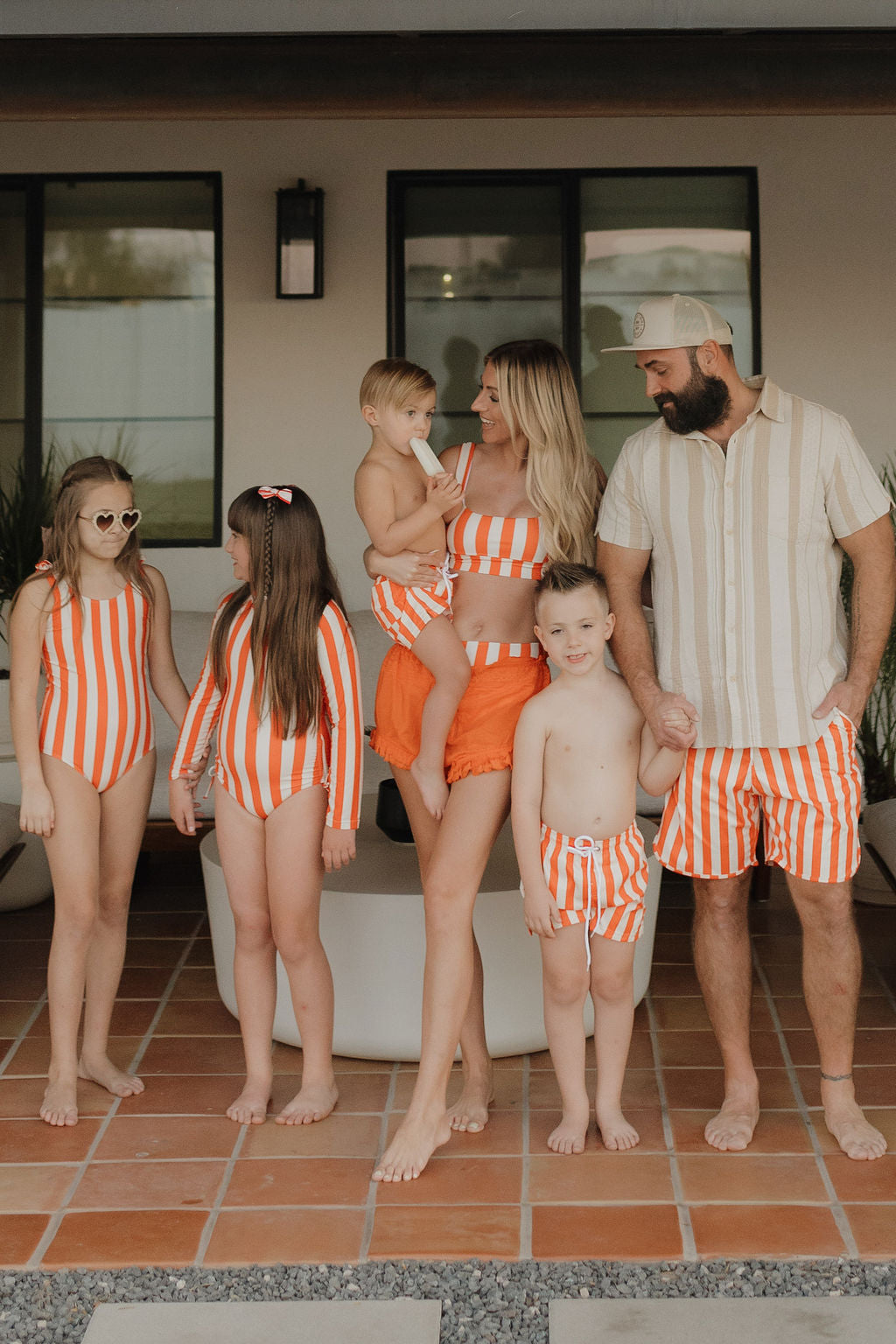 A family of six stands on a tiled patio, wearing matching Poolside swimwear in orange and white stripes. The mother dons a forever french baby Women's Two Piece Swimsuit while holding a toddler. The father holds another child's hand as two girls and a boy smile nearby, all framed by lush plants.