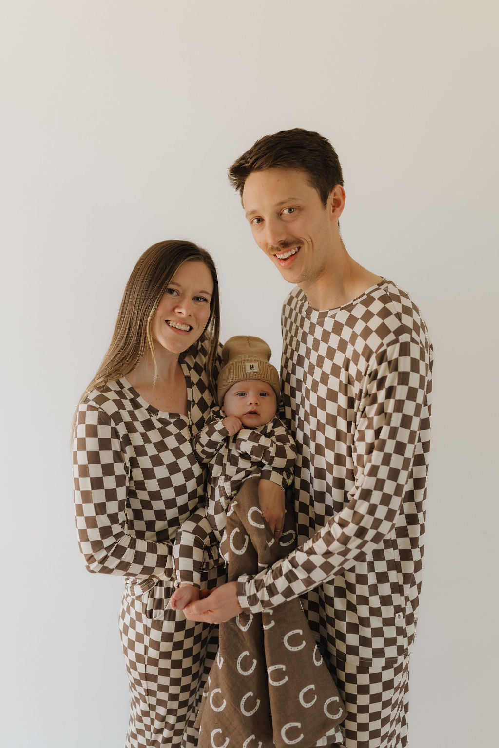 A couple in matching brown and white checkered outfits, embrace their baby wrapped in a bamboo hypoallergenic "C" letter blanket from forever french baby. The baby wears a matching beanie against a plain white backdrop, highlighting the coordinated look and breathable Dutton fabric.