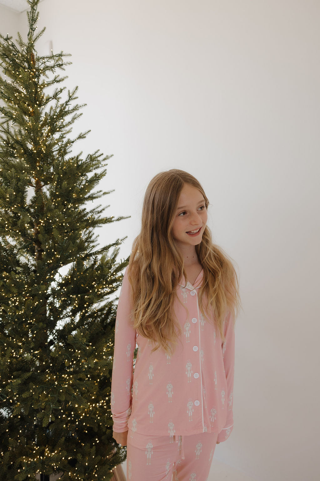 A young girl with long blonde hair, wearing forever french baby Pre-Teen Flare Bamboo Pajamas | Nutcracker, stands smiling by a Christmas tree with white lights in a bright, minimal room.
