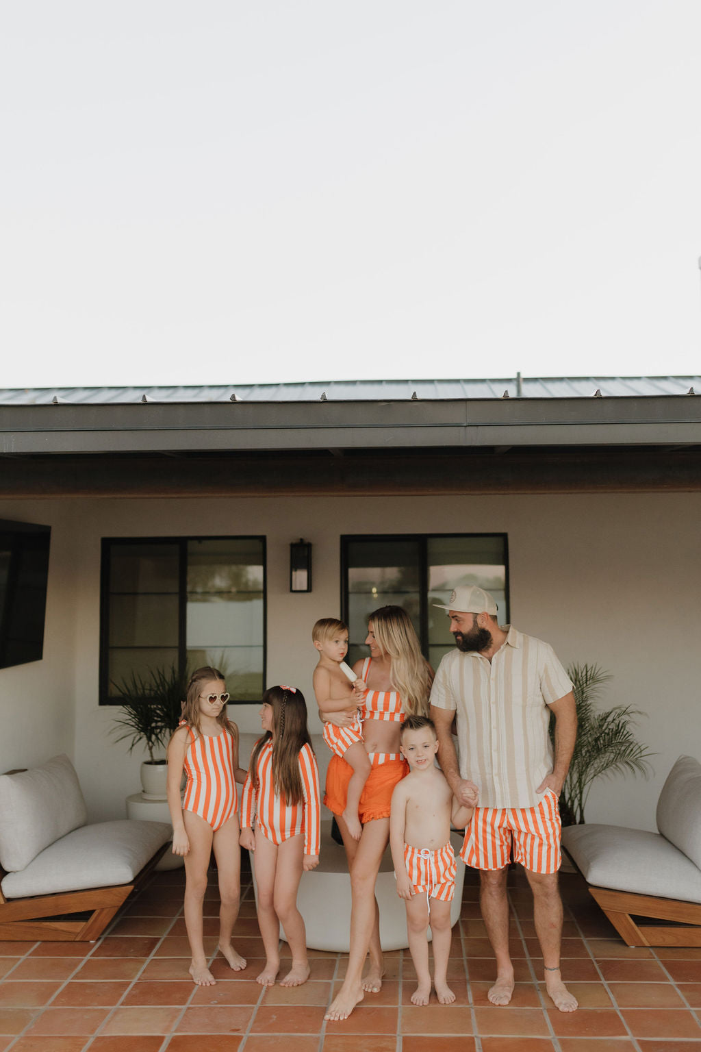 A family of six, wearing matching orange and white swimsuits from the Poolside collection by forever French baby, poses outdoors. The group includes two adults, two girls in Girls Sleeveless Swimsuits, and two boys. They stand on a patio with modern furniture and a metal-roofed building behind them.